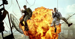 Two men suspended by wires in front of a large orange and yellow explosion.