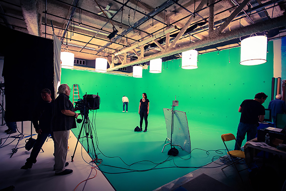Film crew members and equipment in a large studio featuring a bright green screen backdrop.