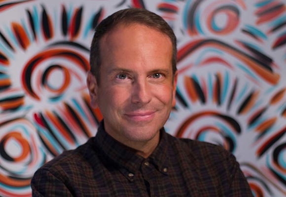 A man with short brown hair and a plaid shirt smiling in front of a colorful abstract pattern.