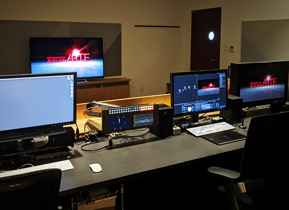 A dark editing studio featuring several computer monitors displaying video production software and logos.