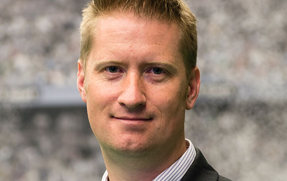 A man with short blond hair and blue eyes smiling slightly against a blurred background.