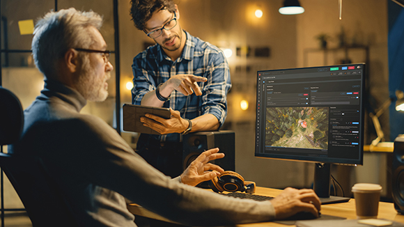 A man sits at a desk using a computer while another man stands beside him pointing at the monitor.