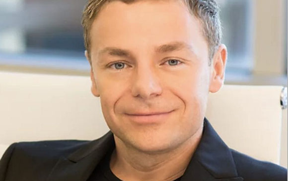 A man with short light brown hair and blue eyes smiling slightly while wearing a black blazer.