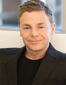 A man with short blond hair and blue eyes smiling slightly, wearing a black blazer and t-shirt.