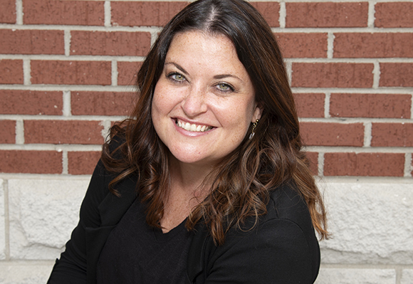 A smiling woman with long dark hair wearing a black top against a red brick wall.