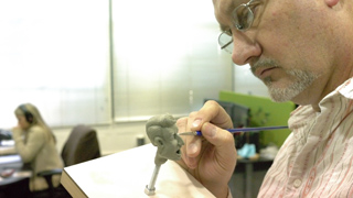 A man in glasses uses a fine tool to sculpt a small clay human head on a workbench.