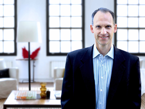 A man wearing a dark blazer and blue striped shirt stands in a bright, modern room with large windows.