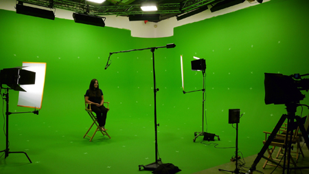 A woman sits in a director's chair in a large green screen studio surrounded by lighting and camera equipment.