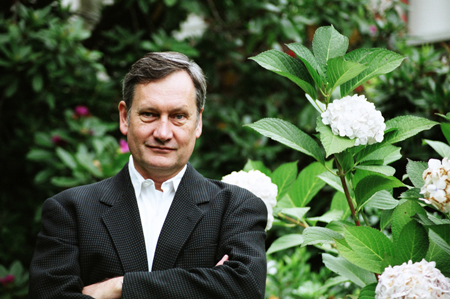 A man in a dark blazer and white shirt stands with arms crossed in a lush garden with white hydrangeas.