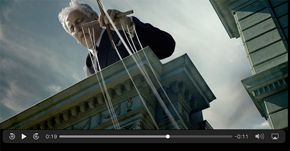 A giant man in a suit pulls white strings from the top of a large stone building against a cloudy sky.