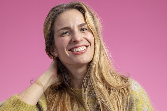A smiling woman with long blonde hair tilts her head while resting her hand on her neck against a pink background.
