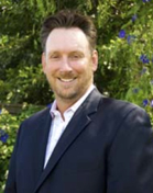 A smiling man with short brown hair wearing a dark blazer and a light-colored button-down shirt.