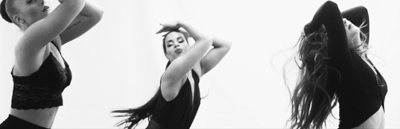 Three women in black attire performing expressive dance movements against a plain white backdrop.