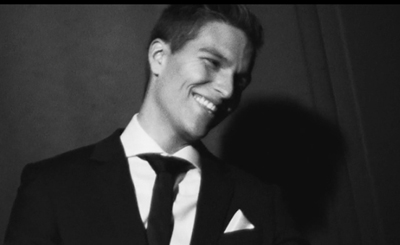 A smiling man in a dark suit and tie looking to the side in black and white.