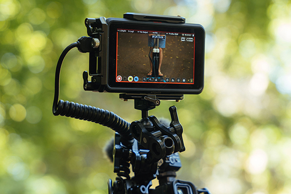 A video camera monitor on a tripod shows a person standing on a dirt path.