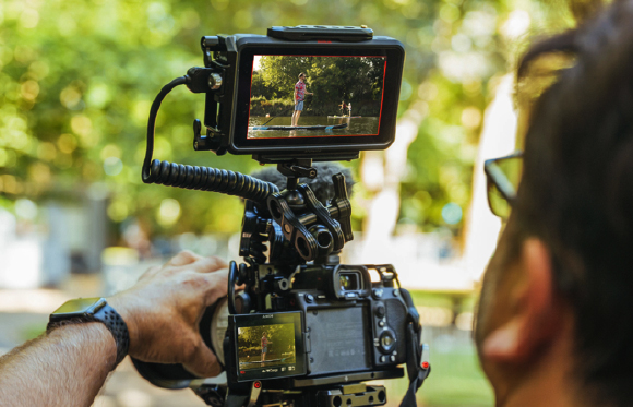 A person uses a professional video camera with an external monitor to film someone in a park.