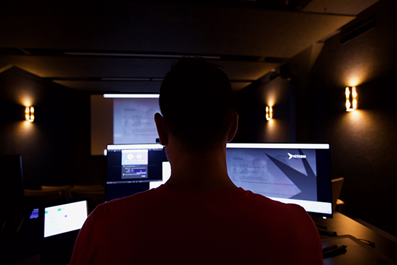Silhouette of a person in a red shirt looking at several glowing computer monitors in a dark room.