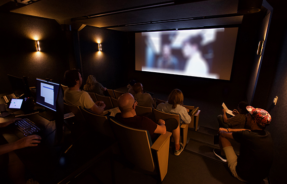 A group of people sit in a dark movie theater watching a film projected on a large screen.