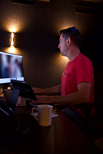 A man in a red t-shirt sits at a desk using a computer and tablet in a dark room.
