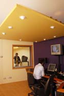 A person in a white shirt sits at a desk in a purple control room looking through a window.