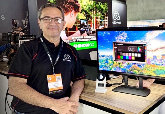A man wearing glasses and a black Atomos polo shirt stands beside a large computer monitor on a wooden desk.
