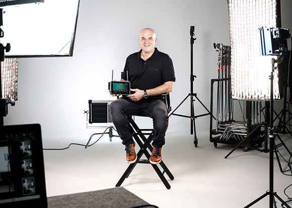 A man in a black polo shirt sits on a stool in a studio, holding a small video monitor.