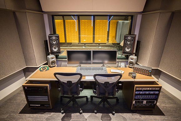 A recording studio desk with dual monitors, studio monitors, a mixing console, and two ergonomic chairs.