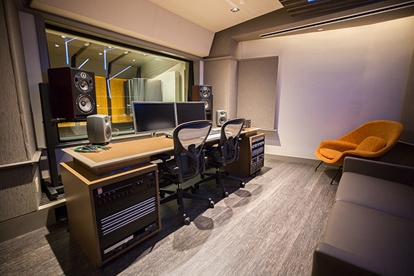 A music studio control room with a large desk, dual monitors, studio monitors, and an orange lounge chair.
