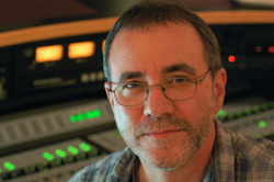A man with short hair and glasses smiling in front of audio mixing equipment.