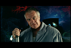 A man in a grey shirt holds a flashlight against a dark, blue-toned background with red branch-like patterns.
