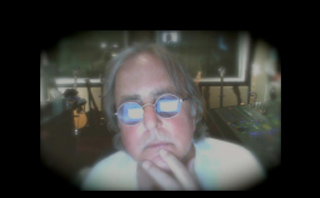 A man with long hair and round glasses looks forward with his hand on his chin.
