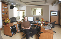 A man sits at a large wooden desk equipped with audio mixing consoles, monitors, and recording equipment.