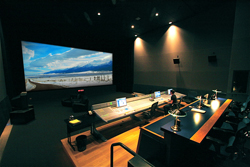 A dark control room featuring a large screen showing a landscape and long technical workstations.
