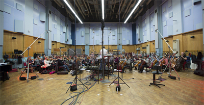 An orchestra performs in a large studio with high ceilings, wooden floors, and various microphones on stands.