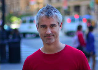 A man with short, spiked gray hair and a red shirt smiles in a blurred city street.