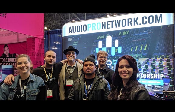 Six smiling people pose for a group photo in front of a large blue and white Audio Pro Network display.