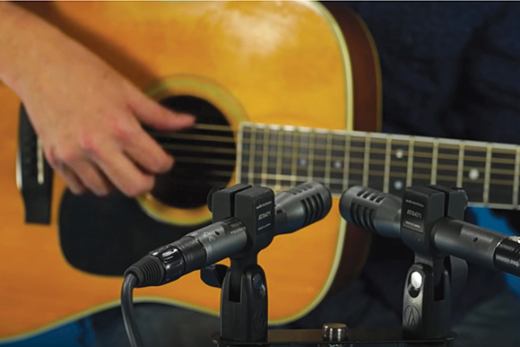 A person's hand strums an acoustic guitar positioned in front of two professional microphones.