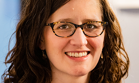 A smiling woman with wavy brown hair and tortoiseshell glasses.