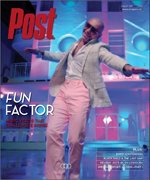 A man in a light suit and pink pants with arms outstretched stands in front of a colorful, modern building.