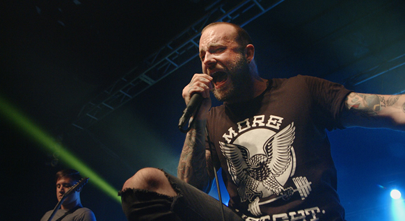 A man with tattoos and a beard sings intensely into a microphone during a live performance.
