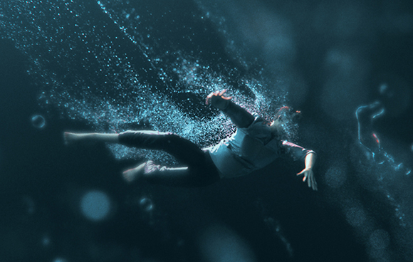 A person in a white shirt and dark pants floats underwater surrounded by rising bubbles.
