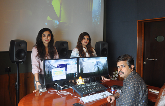 Three people stand behind a desk with dual computer monitors and studio speakers in a dark room.