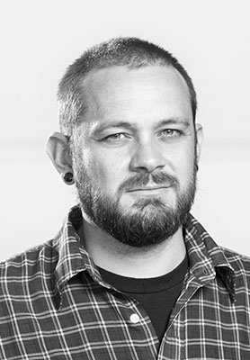 Black and white headshot of a man with a beard, short hair, and an ear gauge wearing a plaid shirt.