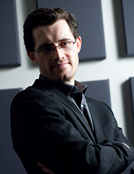 A man with glasses and a dark suit stands with arms crossed against a background of acoustic panels.