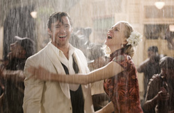 A man in a white suit and a woman in a red patterned dress laugh together while standing in heavy rain.