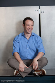 A man with short brown hair, wearing a blue button-down shirt and brown pants, sits cross-legged and laughs.