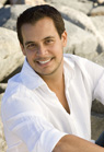 A smiling man with short dark hair wearing a white button-down shirt, sitting against a rocky background.