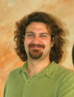 A man with brown curly hair and a goatee wearing a green collared shirt.