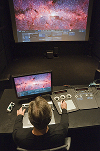 A person sits at a desk with two monitors, editing a colorful nebula image using a control surface.