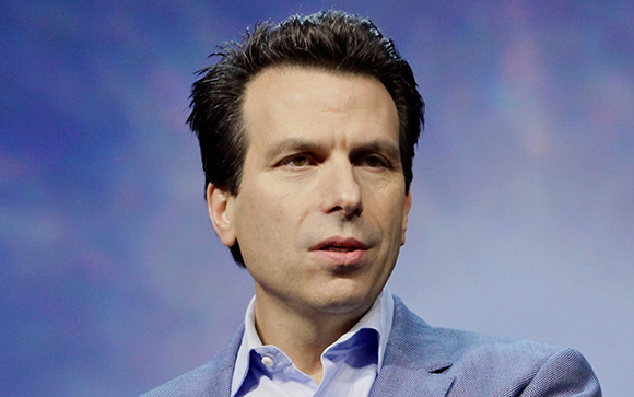 A man with dark hair wearing a light blue suit jacket and a light blue button-down shirt against a blue background.